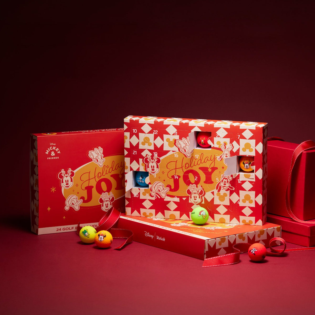 Disney Advent 2025 Holiday Joy boxes in red and white patterned packaging, displayed on a dark red background with small colorful advent balls in front.