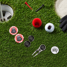 A set of Star Wars Darth Vader themed golf accessories arranged on a green grass background, including a red golf ball, a white golf ball, two metal chips, a metal divot tool, a metal flag, and a black glove.